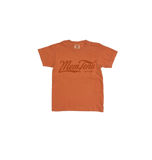 An orange Comfort Colors t-shirt with "Youth Memphis Tenn Cursive on Terracotta" printed in the center, hanging on a white hanger against a striped background.
