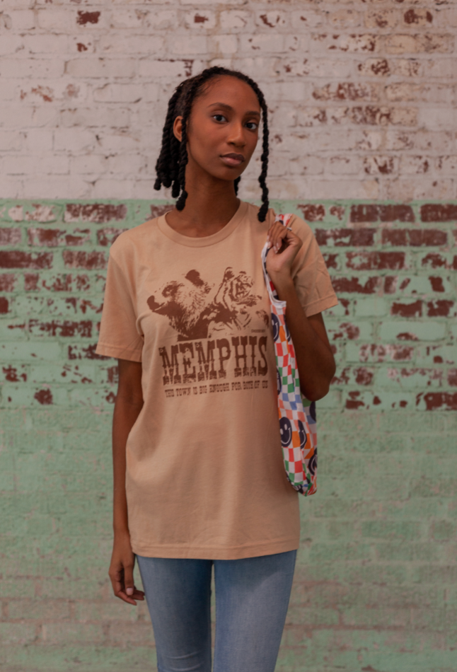 A person stands against a weathered brick wall, wearing "The Both of Us" Short Sleeve Tee by Choose901 Merch Shop and holding a multicolored bag, showcasing the Tigers' fierce spirit.
