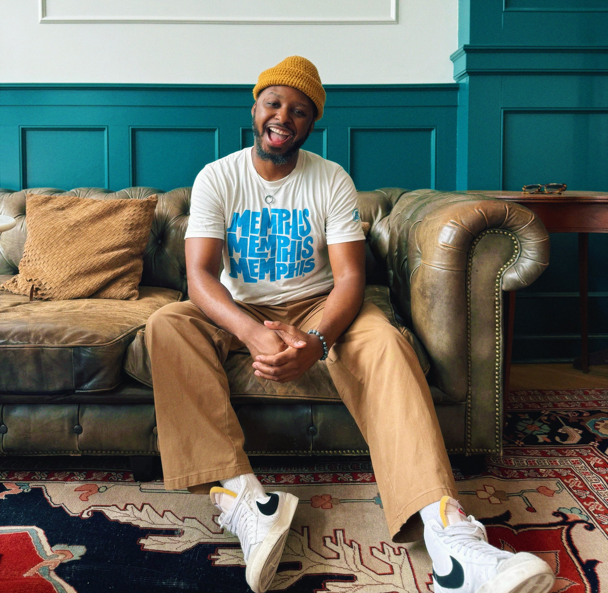 A man in a yellow beanie, tan pants, and white sneakers smiles while wearing a white "Memphis, Memphis, Memphis by Eso Tolson" tee from Choose901 Merch Shop, seated on a brown leather sofa against teal paneled walls.