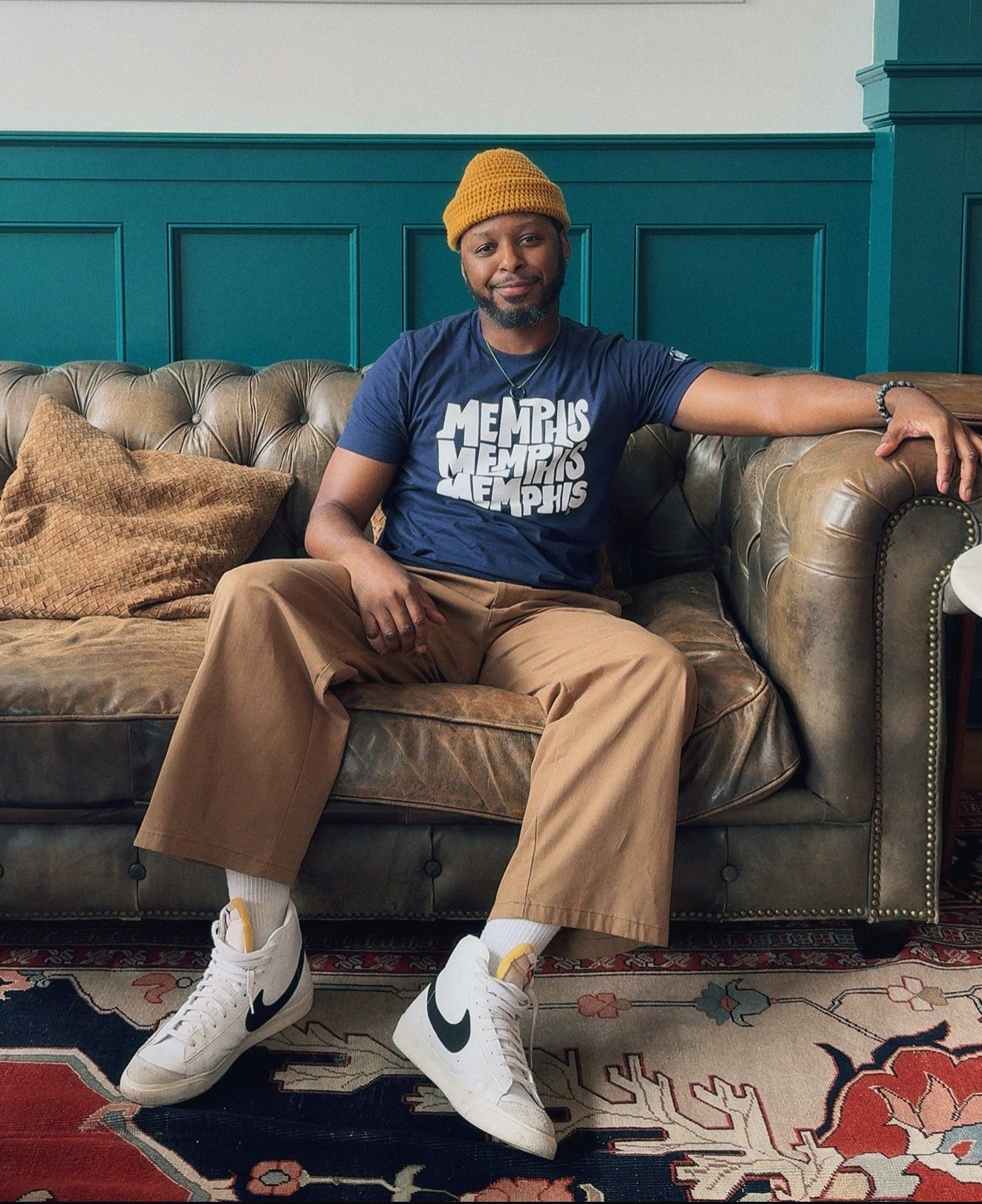 A man wearing a yellow beanie, tan pants, and white sneakers sits on a brown leather couch atop a patterned rug. He sports a blue "Memphis, Memphis, Memphis by Eso Tolson" tee from Choose901 Merch Shop.