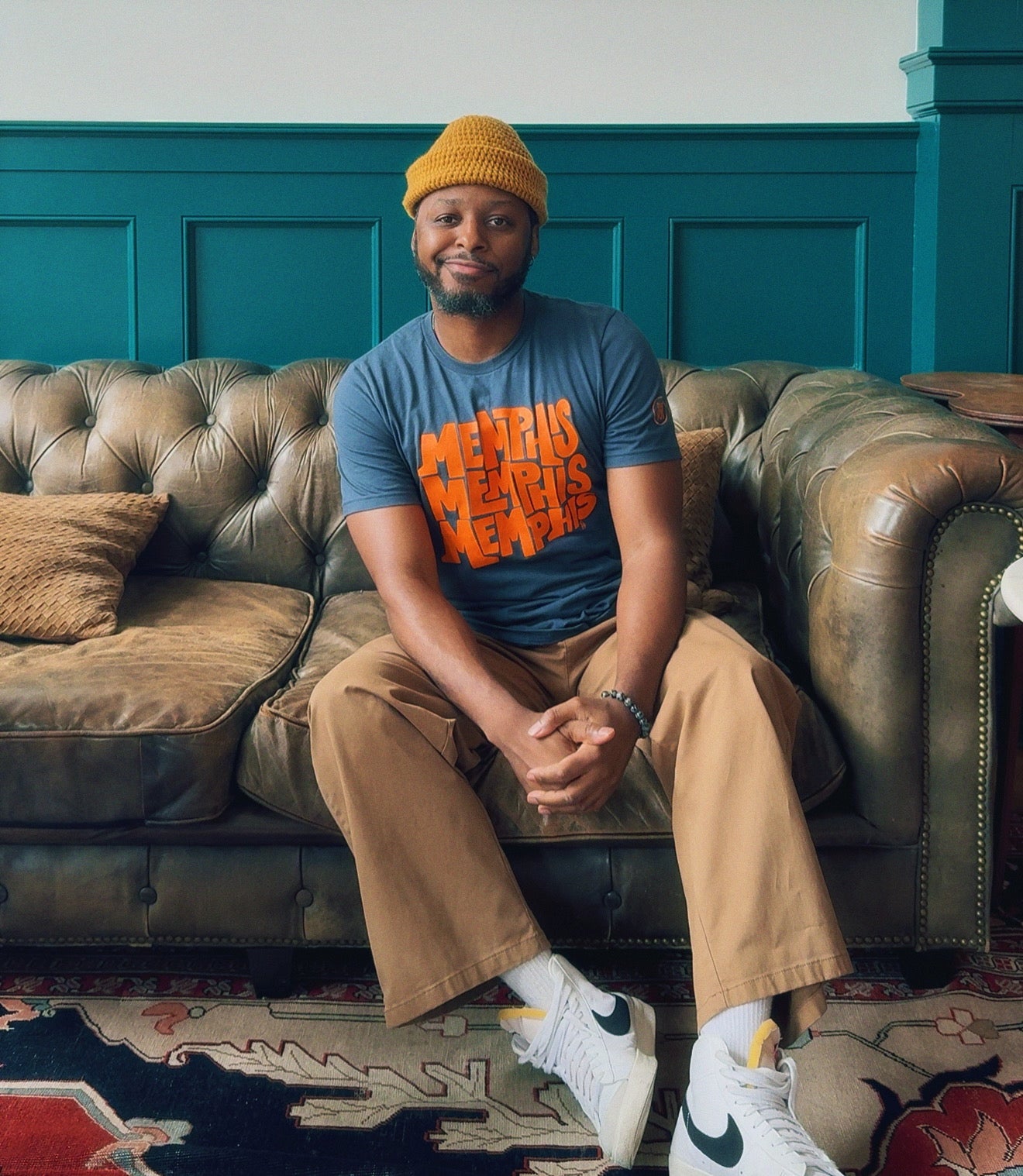 A man in a yellow beanie, tan pants, and white sneakers wears a blue "Memphis, Memphis, Memphis by Eso Tolson" tee from Choose901 Merch Shop while sitting on a leather sofa with green paneling and a patterned rug behind him.