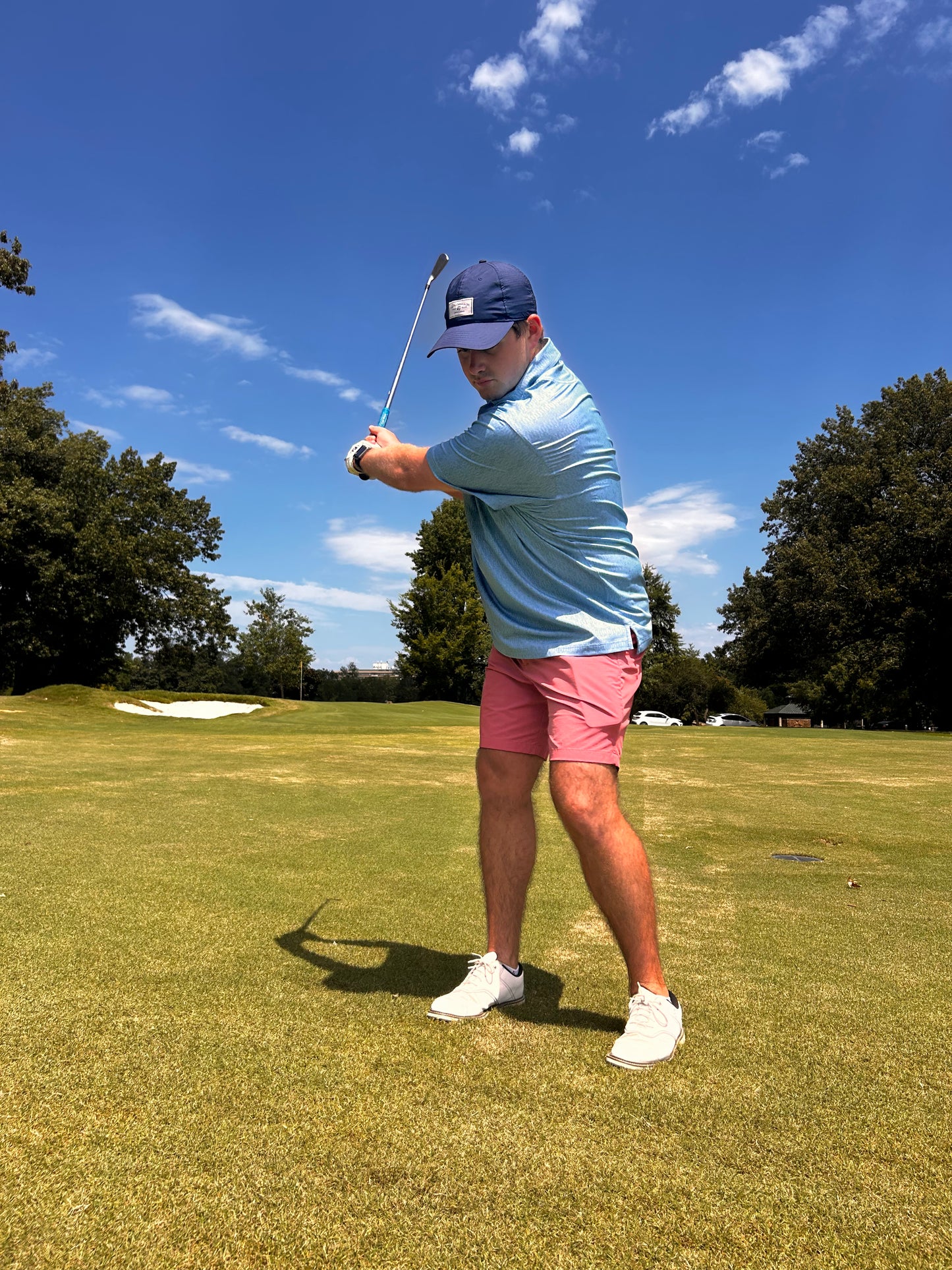 A person wearing The Kings' Guitar Polo from Choose901 Merch Shop, complemented by pink shorts and a blue cap, is holding a golf club in a mid-swing position on a golf course under a clear, sunny sky.