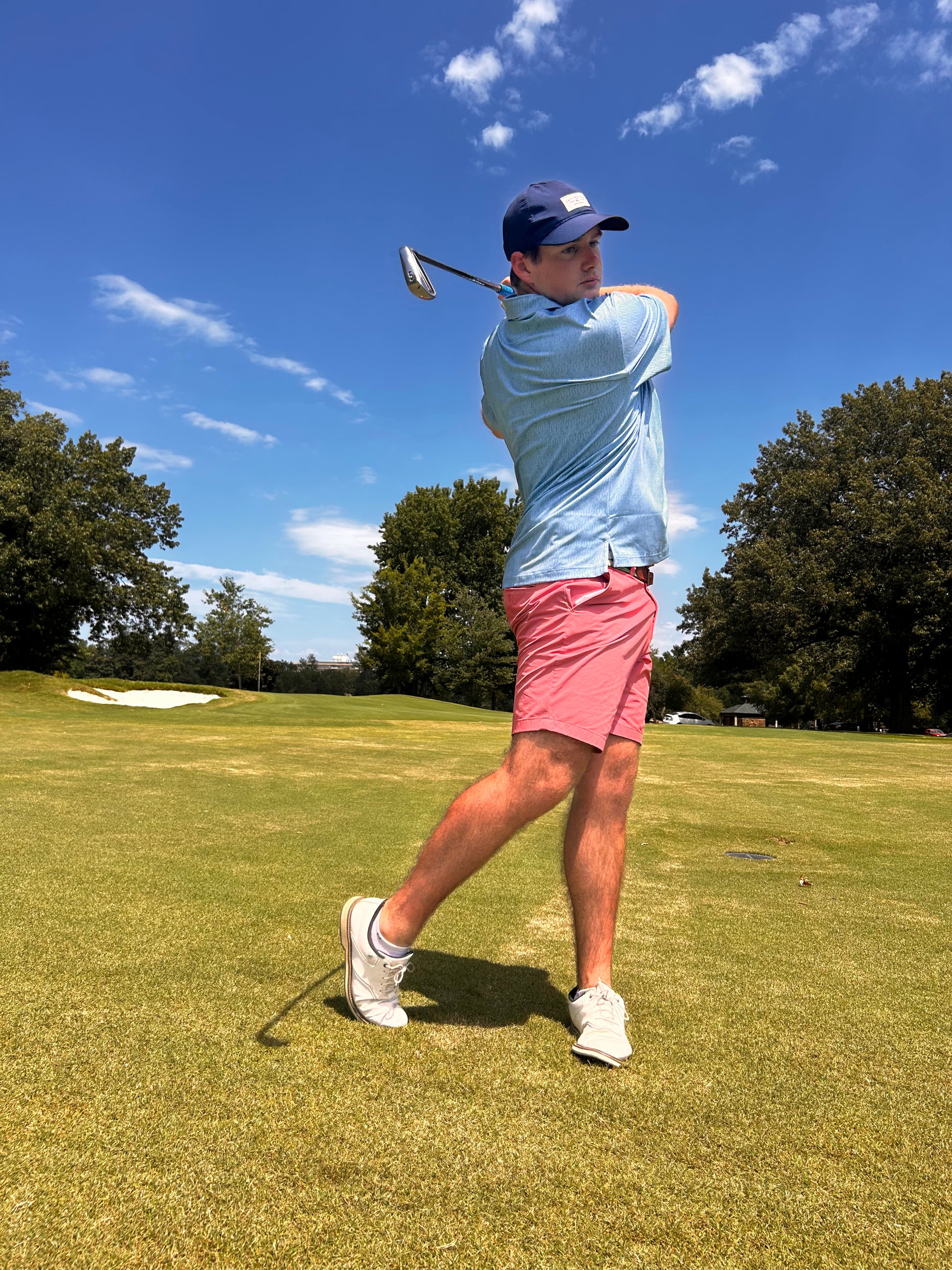 A man in The Kings' Guitar Polo from the Choose901 Merch Shop, paired with pink shorts and a blue cap, swings a golf club on a grassy golf course under a partly cloudy sky. Trees and a sand trap are visible in the background.