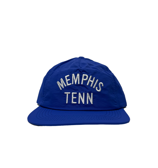A Memphis Tenn Nylon Hat Royal Blue with "memphis tenn" embroidered in white, placed on a striped gray background by Choose901 Merch Shop.