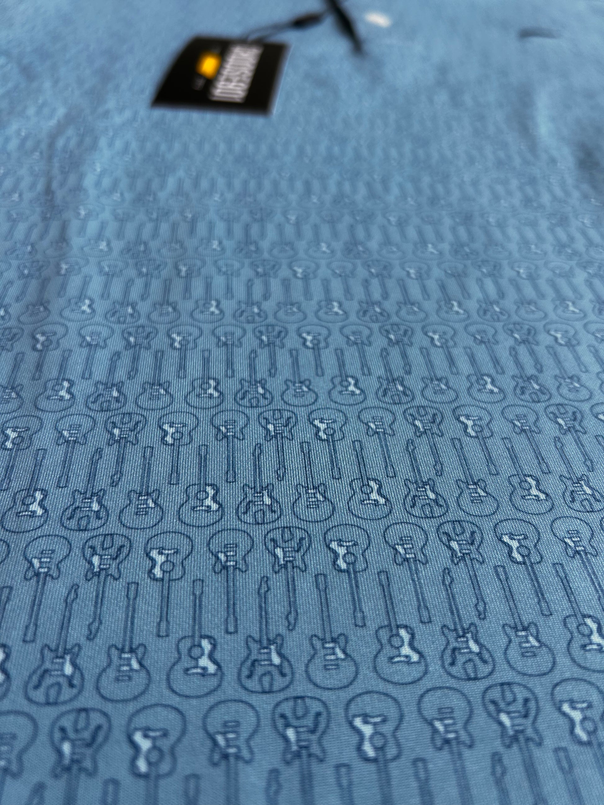 Close-up of The Kings' Guitar Polo, a comfortable shirt in blue fabric with a repeating pattern of small guitars. A portion of a black clothing label from Choose901 Merch Shop, featuring text in white and yellow, is visible near the top of the image, highlighting this versatile polo's unique design.