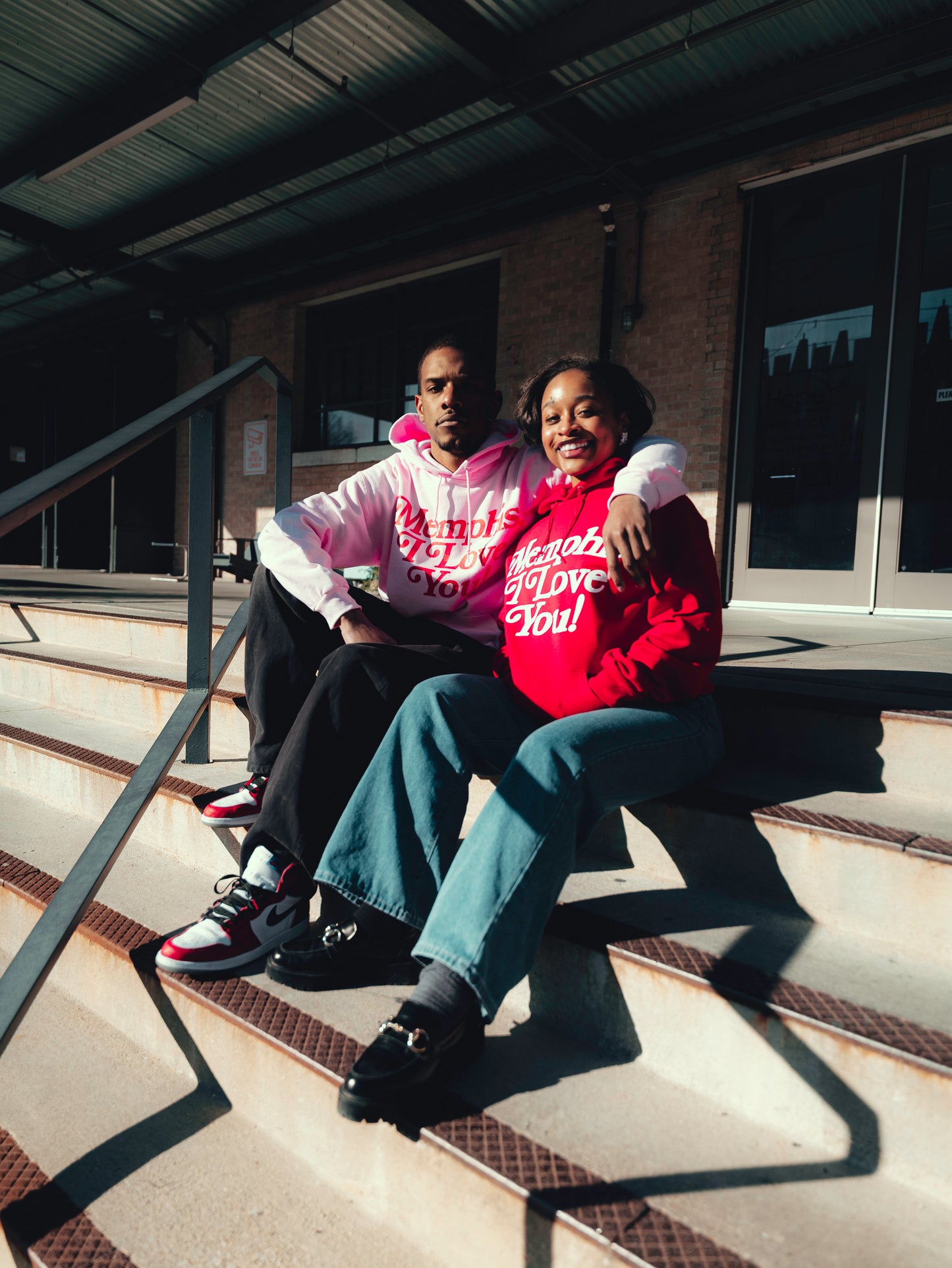 Two people sit on outdoor steps in Memphis, joyfully smiling at the camera, donning Choose901 Merch Shop's Valentine's edition "Memphis I Love You" hoodies with script text—one in a white hoodie and the other in red.