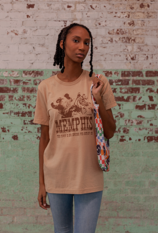 A person stands against a brick wall, wearing a dazzling "The Both of Us" tee from Choose901 Merch Shop and jeans, holding a colorful patterned bag.