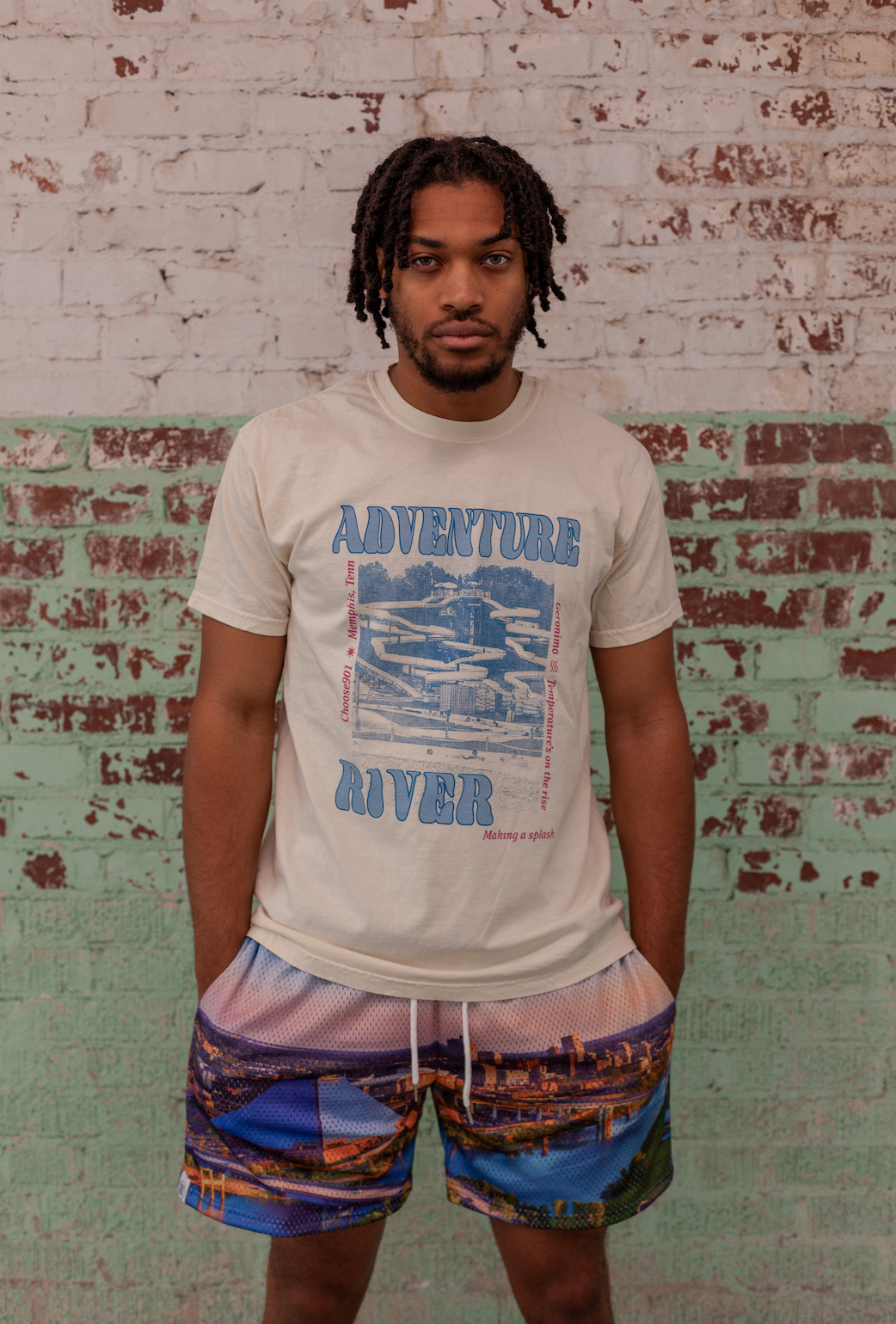 Man standing in front of a brick wall wearing a Adventure River Shirt Ivory from the Choose901 Merch Shop and Memphis patterned shorts.