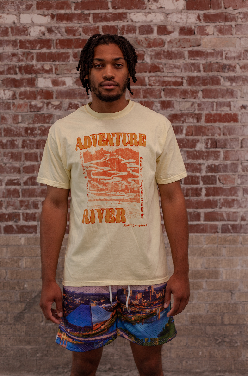 A young man in a Choose901 Adventure River Shirt Yellow and patterned shorts standing against a brick wall in Memphis.